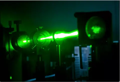 STFC A bright, glowing green laser beam shoots horizontally through a dark laboratory. It passes through circular metal instruments and lenses, getting wider and more intense. The surrounding equipment is mostly in shadow, so the vivid green light is the dominant, almost otherworldly feature against the black background.