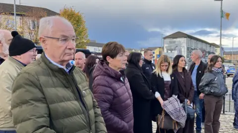Shows about a dozen people standing around with a mural in the background and a flagpost with an Irish tricolour on it