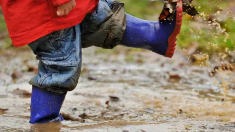 Getty Images The bottom half of a toddler in blue wellies and red coat, splashing in a deep, muddy puddle.