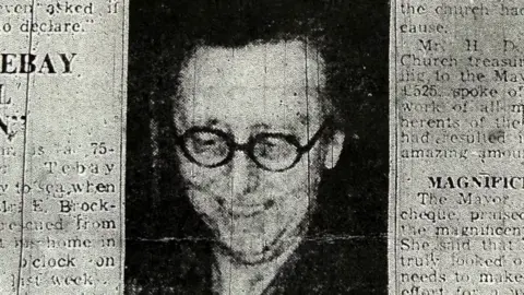 Elsie Stannard photographed black and white wearing round spectacles looking down smiling. The photograph is from a scanned copy of the Westmoreland Gazette. 