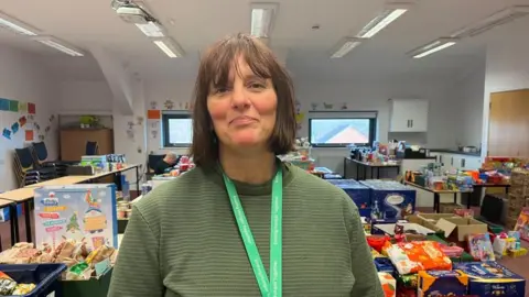 ELLEN KNIGHT/BBC Liz pictured looking into the camera and smiling. She's wearing a light green jumper, with a green Trussell Trust lanyard. She's stood inside in a large, bright room, with five rows of tables behind her. On the tables are crates filled with food - nuts, pickles, chocolates, crackers and more - toys, and toiletries.