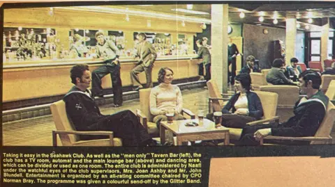 Royal Navy/Navy News A grainy photo in Navy News which shows the Seahawk Club in 1975. There is a long bar along the back wall which three men are leaning against. There are low coffee tables with cushioned armchairs. In the foreground there are two women and two men in sailor suits sitting with drinks in front of them.
There is a caption which begins: Taking it easy in the Seahawk Club and goes on to describe the club.