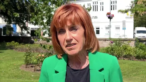 BBC Woman with red hair, black top and green jacket standing in front of a park and a white building.
