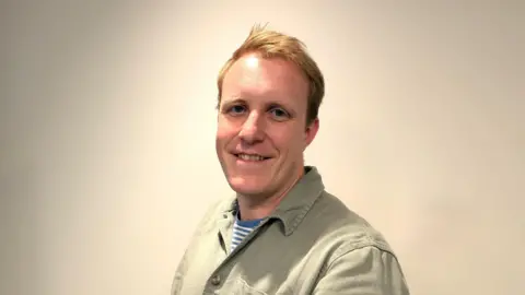 Steffan Evans A young man with short blond hair, smiling and wearing a light brown shirt.