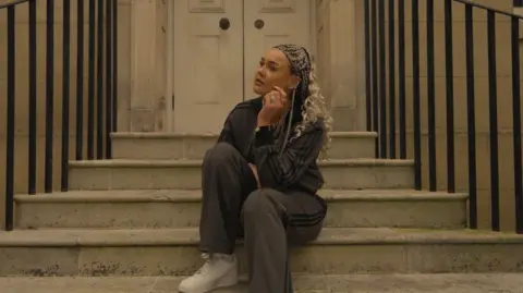 Harriet Laws The photo shows Harriet Laws sitting outside on the steps leading up to a doorway. She is looking to the side, wearing a grey Adidas tracksuit and gold necklaces.