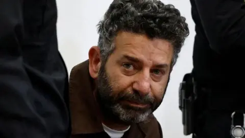 Close up of Saif Abu Keshek sitting accompanied by two standing Israeli police officers at a court in Israel. He has short, greying hair and a beard.