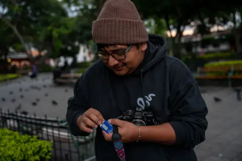 Fritz Pinnow Ronald Ottoniel, donned in a knit cap and thick-framed glasses, inserts his new film roll into his camera in a square located in Antigua. Pigeons can be observed on the ground in the backdrop.