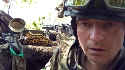 Sergiy Sylantyev, a man in Ukraine's military uniform, with a heavy machine gun and ammunition, taking a selfie.