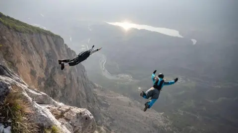Getty Images Two people in colourful suits with parachute packs on their backs jump from a cliff.  Beyond them is a valley with a green floor and the sun shining off a lake.