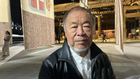 Photograph of the artist Ai Weiwei. He is pictured alongside his artwork in the giant warehouse space at Manchester's Aviva Studios. 