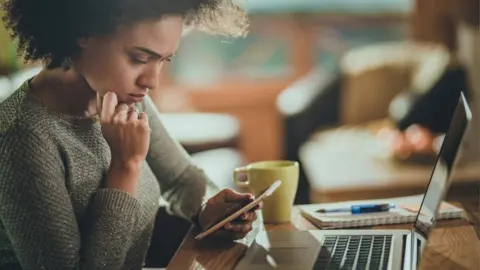 Getty Images A woman holds a phone while sat at a desk