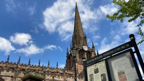 Shot of Rotherham Minster alongside a sign which says Rotherham Parish Church on it. Picture taken on a sunny, blue sky day.