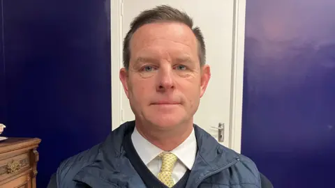 BBC A man with short brown hair wears a navy v-neck sweater, navy gillet, white shirt and yellow tie. Behind him is a white door which splits a purple wall. 