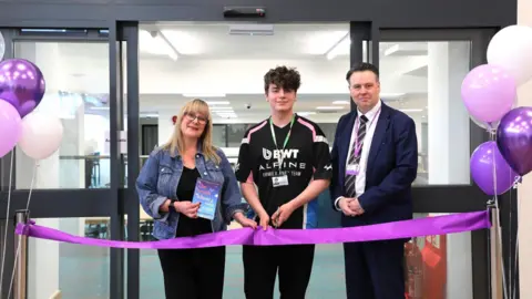 A woman and two men standing in front of a purple ribbon, with purple balloons at either end in front of the glass doors of the library. The woman has blonde hair and is holding a blue book. The man in the middle has brown curly hair and is holding scissors to the ribbon. On the right the man has dark hair and is wearing a suit