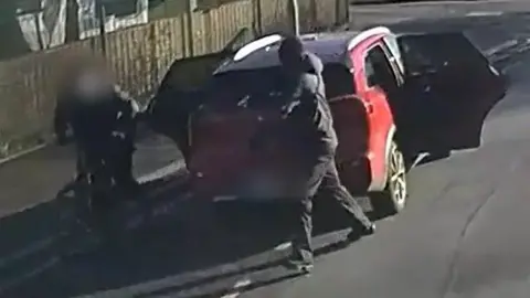 GMP A man in black clothing and a black head covering points a silver handgun at another man, also in black, who is attempting to ride away on an electric bike. They are standing in front of a red taxi with its doors open. 