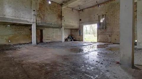 Moveli The interiors of the power house. There are puddles on the floor, floor to ceiling beams and a pile of rubbish in the furthest corner.