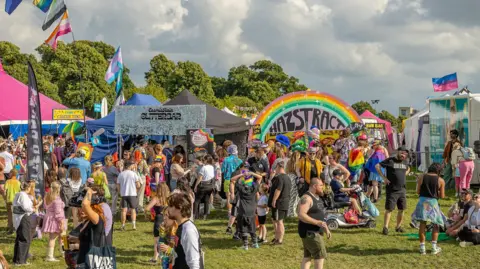 Bristol Pride / Dan Weill Photography Crowds walking and checking out a number of stalls on the grassy Downs during Bristol Pride in 2024. The weather is sunny, with some clouds in the sky.