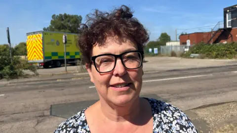 Julie, a woman with short curly hair. she is wearing a floral blue, black and white top, smiling for the camera. Julie is standing on the road, with an ambulance in the background.