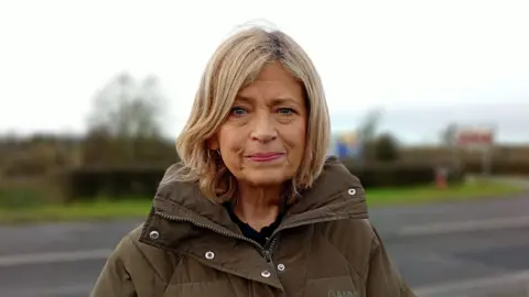 Diana looking at the camera. She stands at the side of the road wearing a green puffer coat. She has shoulder length blonde hair and is wearing gold earrings.