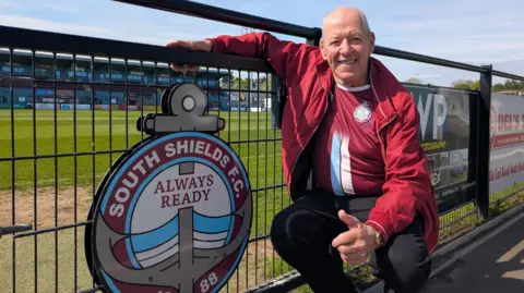 Jim Taroni, an 80-year-old bald man, smiles warmly as he crouches by a South Shields FC crest attached to the fence surrounding the vibrant grass pitch. He's wearing a red bomber jacket over his South Shields home shirt.