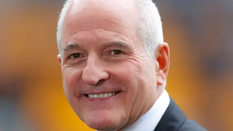 A man with white hair is smiling and looking at the camera. He is wearing a black jacket and white shirt and the background is blurred.