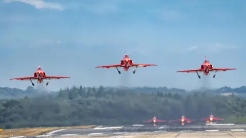 Hang Ross Three red jet planes take-off simultaneously with vapour trails behind them. Through those trails the image of the next three planes waiting can be seen on the runway behind them.