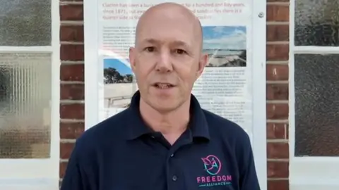 Freedom Alliance Martin Davies stands outside Clacton station. He is wearing a dark blue t shirt with the Freedom Alliance logo on.