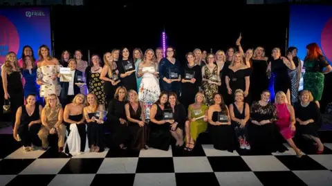 Bagnalls A large group of people posing together on a black-and-white checkered floor at an indoor event. Many individuals are dressed in formal or semi-formal attire, including dresses and evening wear. Several people are holding rectangular awards or plaques.