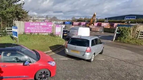 Google Entrance to Sixfields recycling centre showing a purple Sixfields HWRC sign on a fence to the left, and a queue of cars and vans heading through the gate.  There is a yellow digger in the background and a row of shipping containers.