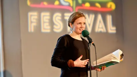 McNish stands at a mic with a book in her hand in front of a sign saying Festival