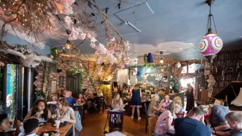 The Steel Cauldron There are several adults and children sitting at tables in a cafe, which is decorated with branches of blossom, trees, fairy lights and colourful lamps
