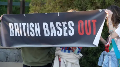 Jessica Parker/BBC Three people standing behind a black sign they are holding up that reads white and red: "British Bases Out."