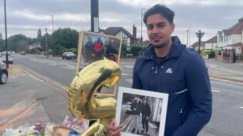 A man with brown hair wearing a blue zip top stands in front of a pile of flowers, balloons and photos. He is next to a main road in a residential area and is holding a white framed photo of two men.