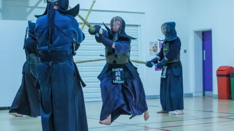 Andy Lomas Chloe mid-fight while training. She is wearing her full dark blue Kendo uniform and caged helmet.