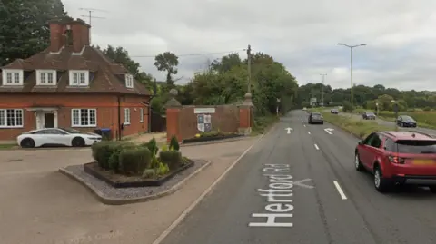 Google A dual carriageway which runs directly outside the entrance of a school drive which has some small hedges and a building at the end of it.