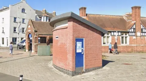 The picture shows a small brick building with a curved roof and a blue door sitting in an open paved area. It looks like an old public toilet block. Behind it there are older red‑brick buildings with tall chimneys and black‑and‑white timber detailing, along with a larger pale-coloured building on the left. A few people are walking across the open space, and the scene is bright with clear blue sky.