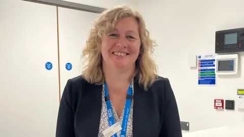 Andrew Turner/BBC Jo Segasby in a white clinical room. She is looking directly at the camera and is smiling. She is wearing a patterned white blouse, navy blazer and has a blue NHS lanyard round her neck.