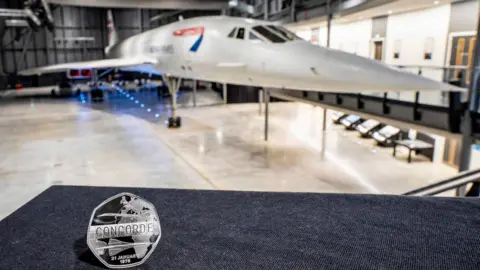The close up of the coin on a blue surface. The reverse or 'tails' side of the coin portrays the aircraft in flight, against a backdrop of a split-flap departure board, with the word Concorde appearing across the centre. The Concorde plane is in the background.