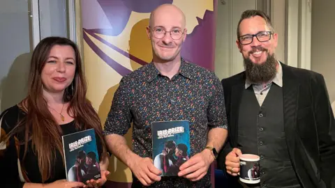 Three people stand together, smiling and holding a copy of the book featuring Tom Paul Martin's new film.