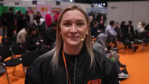 Suz Bannister, a woman with blonde hair wearing a black jacket looks at the camera. She is wearing a microphone.