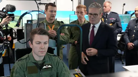 Sir Keir Starmer stood talking to a man operating a flight simulator. He is wearing a dark suit while the young man operating the simulator is wearing a green RAF uniform.