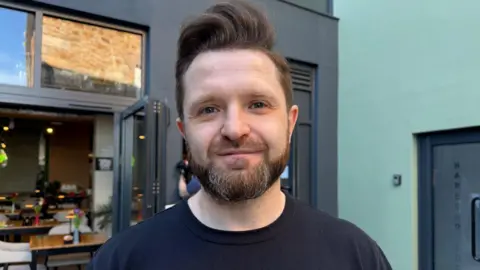 Sebastian Krzeminski is pictured in front of the Gallery cafe which he runs with his wife. He is wearing a black t-shirt and has a beard and brown hair.