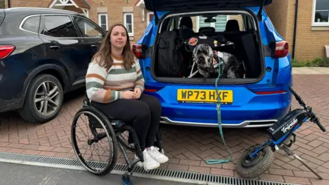 Kay Millar is sitting in a wheel chair next to a blue car with the boot open. Her dog is standing in the boot.