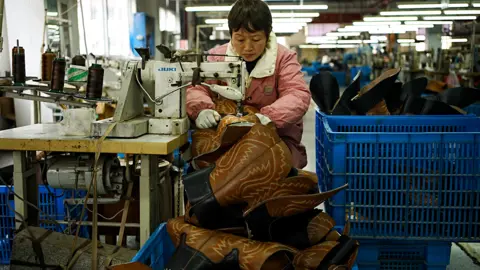 Xiqing Wang/ BBC A worker at the factory sits at a wooden bench with an old sewing machine on it, sewing brown leather cowboy boots. She is wearing a pink and white jacket, and there are large blue containers next to her full of boots.