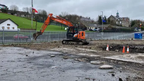 Shows a diggers on a derelect stie with railings on the perimeter and a church and houses in the background