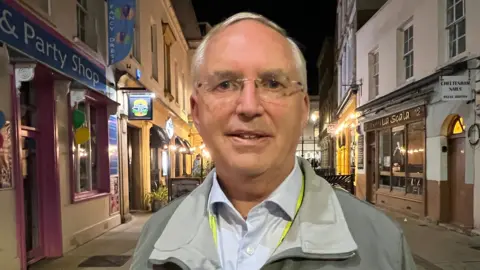 Chris Nelson stands on Regent Street in Cheltenham at night, in the middle of the street, with a party shop and The Beefy Boys to the left of him and La Scala and Cheltenham Nails to the right of him. He is an older man with grey hair, and is wearing thin-rimmed glasses, a grey jacket, a white shirt, and a bright yellow lanyard.