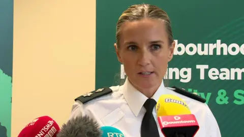 BBC PSNI Ch Supt Gillian Kearney at the press conference. She has blonde hair, pulled back in a middle parting. She wears a PSNI uniform and stands in front of press mics from various outlets across NI and ROI.