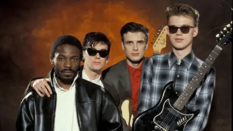 Band Orange Juice - Zeke Manyika (mengenakan jaket kulit hitam dan kemeja putih), James Kirk (mengenakan nuansa) David McClymont (jas abu -abu dan jaket merah) dan Edwyn Collins (kemeja yang tidak sesuai, memegang gitar hitam) terlihat ke kamera