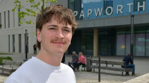 George Collyer is standing on the left. He has short brown hair and a moustache. He is wearing a white T-shirt and is standing in front of the Royal Papworth Hospital which has a large sign above the door that says PAPWORTH.