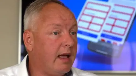 Chris Bee fotografiado en primer plano. Tiene el rostro bronceado, viste una camisa blanca y tiene el pelo corto y canoso. Se puede ver una valla publicitaria al fondo.